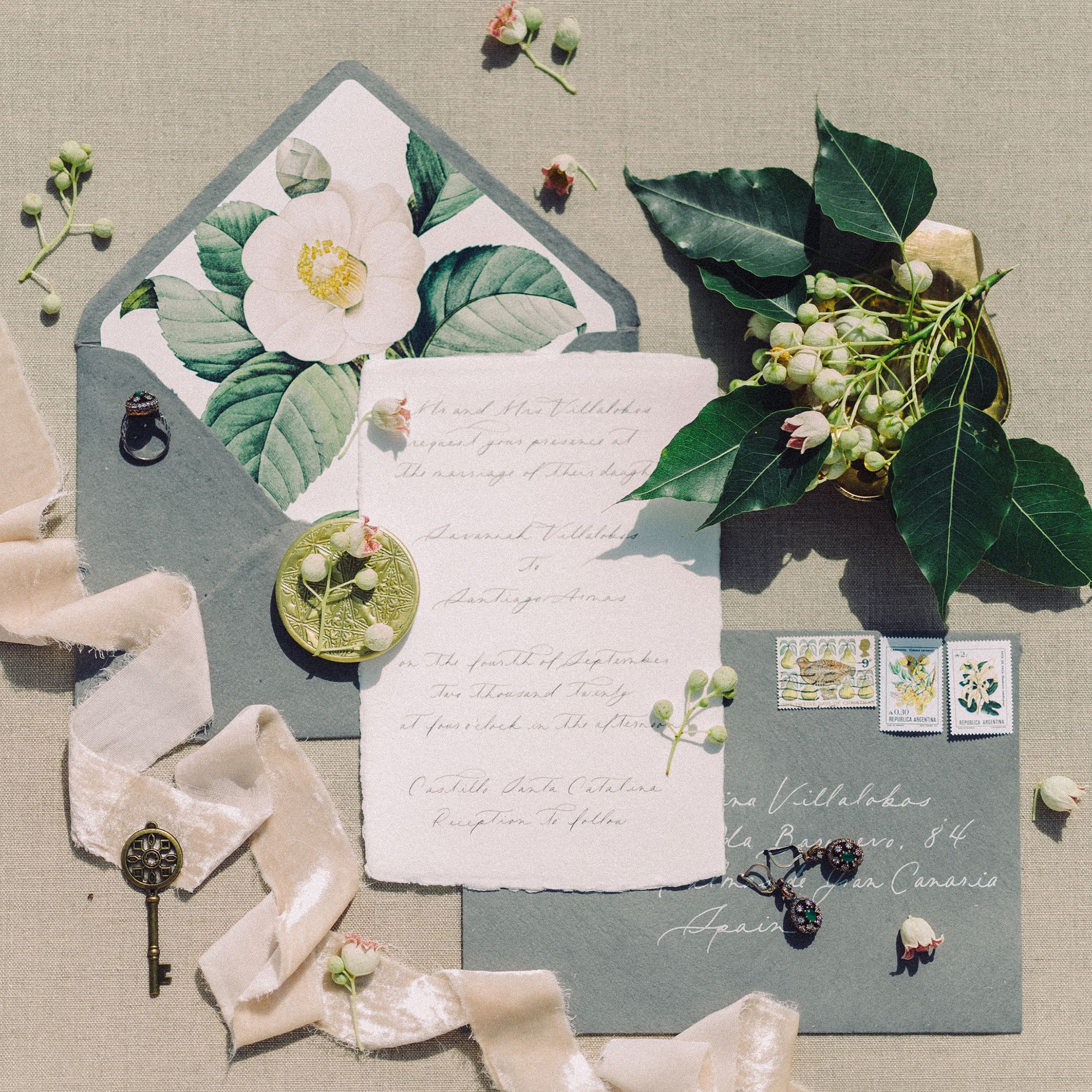 Vintage-inspired wedding invitation suite with sage envelope, camellia botanical liner, wax seal, and antique key on neutral linen backdrop